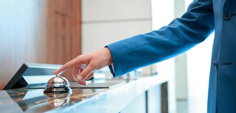 Person im Anzug in einem Hotel, vor dem Empfang, drückt auf eine Tischglocke bezugnehmend auf das GastroSoft Hotel Add-On
